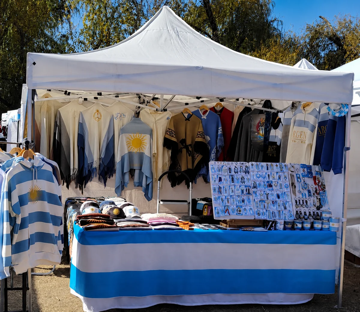 Stand de Argentina La Linda en la feria: ponchos, sweaters y camisetas con el sol de mayo colgados, y escarapelas sobre la mesa